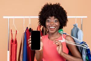 Mobile Shopping Application. Joyful African Female Showing Smartphone Empty Screen To Camera Recomending Fashion And Style Assistant App Posing Near Clothing Rack Over Yellow Studio Background. Mockup photo