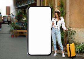 A traveler stands beside a large blank phone billboard while looking at her phone. She has a suitcase next to her and is dressed casually in a stylish outfit, ready for her journey. photo