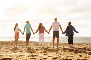 A group of five friends stands together on a sandy beach, holding hands as they face the ocean. The sun sets in the background, creating a warm and inviting atmosphere. photo