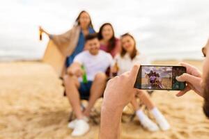 A group of friends poses joyfully on the beach, holding drinks and smiling as one of them takes a picture with a smartphone during sunset. The atmosphere is filled with laughter and happiness. photo