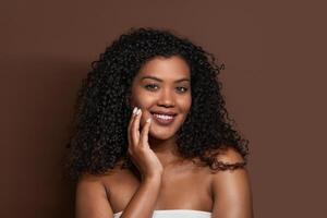 A young woman with curly hair is smiling confidently while resting her hand on her face. She is dressed in a simple top, creating a calming atmosphere with a rich brown backdrop. photo