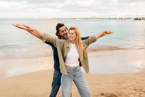 A man and woman share a joyful experience on the beach, with the woman laughing and spreading her arms wide while the man supports her from behind, set against a serene ocean backdrop. photo