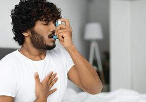 Indian guy suffering from asthma, using inhaler in bed, side view, copy space. Young hindu man woke up with asthma attack, touching chest and using nebulizer, bedroom interior photo