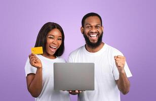 Yes, Huge Sales In Webstore. Excited black couple shopping online, holding credit card, using laptop on blue studio background. African American man making winner gesture, shaking clenched fist photo