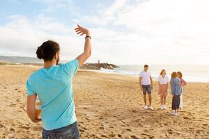 A group of friends stands on the sand at the beach, engaging in conversation while one person waves enthusiastically from nearby. The setting features gentle waves and a distant lighthouse. photo