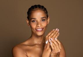 Happy black middle aged woman using hand cream after shower, beautiful female applying moisturizing or nourishing cream on her hands, smiling at camera over brown studio background photo