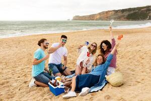 A group of five friends is gathered on the sandy beach, having a cheerful picnic. They each hold drinks and snacks, enjoying the ocean breeze and each other's company under a bright sky. photo
