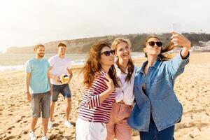 Four friends are having a great time at the beach, taking selfies while two others play volleyball in the background. The sun is shining beautifully over the sandy shore. photo