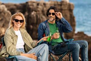 A man and a woman sit on folding chairs near rocky coastline. They are smiling and holding green bottles, enjoying a sunny day by the ocean with gentle waves in the background. photo