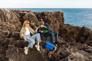 Two friends are sitting on rocky terrain near the ocean, enjoying snacks and drinks in portable chairs. The sun shines brightly as they share a light moment together. photo