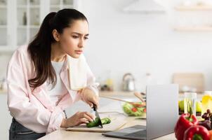 Portrait of serious focused young lady cooking fresh organic salad at home in modern kitchen, using and looking at laptop on countertop, looking closer at screen, reading recipe, free copy space photo