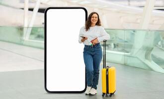 A young woman stands confidently next to a large blank mobile screen, holding a smartphone and travel documents while her yellow suitcase rests beside her in a contemporary terminal. photo
