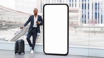 A smartly dressed traveler black guy stands beside a large blank screen at a contemporary airport, with a suitcase in tow, while checking his smartphone, suggesting he is preparing for a journey. photo