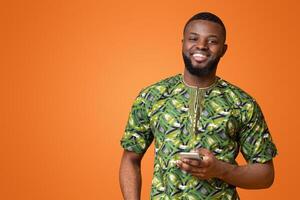 Smiling black guy in authentic african costume using mobile phone, yellow background, copy space photo
