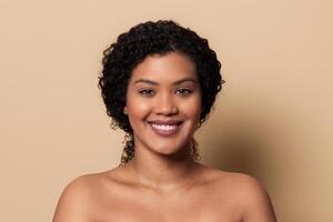 A woman with natural curly hair smiles warmly against a soft beige background. Her bright expression conveys confidence and joy, emphasizing her beauty and poise in a simple setting. photo