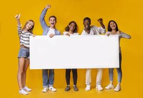 Happy smiling group of multiracial students standing together and displaying white blank placard, yellow studio background photo