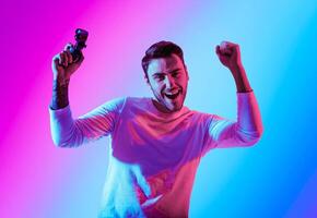 Winning in computer game. Happy man raises hands with joystick, celebrating success, in neon, studio shot photo
