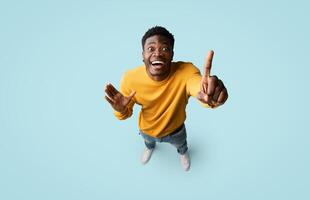 Top view of inspired creative african american millennial guy in casual outfit pointing finger up over blue studio background, having nice idea, showing eureka gesture, full length, copy space photo