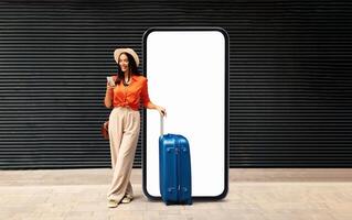 A woman stands confidently with a suitcase and coffee, wearing a fashionable outfit. She enjoys the outdoor atmosphere next to a large blank screen, perfect for ads or displays. photo