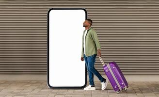 A young black man strolls through a modern space, pulling a purple suitcase behind him. He walks beside a towering blank smartphone display, suggesting travel and technology. photo