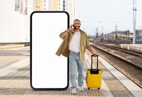 A man stands at a train station, holding a suitcase while talking on his phone. Beside him is an oversized smartphone, emphasizing modern travel and connectivity. photo