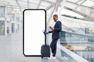 African American businessman stands casually in an airport terminal, holding a smartphone while resting a suitcase nearby. The terminal features modern architecture and a blank digital display. photo