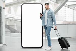 A traveler waits at a modern transit hub, checking a smartphone while standing beside a large blank billboard. The area features sleek architecture and a contemporary design. photo