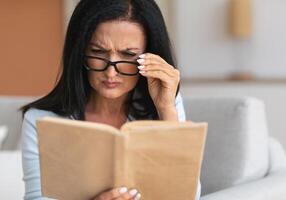Portrait of confused senior woman squinting to see more clearly, wearing and touching eyeglasses, trying to read paper book, having difficulties seeing text because of bad vision problems and issues photo