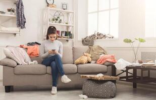 Desperate helpless woman sitting on sofa in messy living room. and chatting on mobile, surrounded by many stack of clothes. Disorder and mess at home, copy space photo