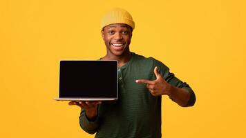 Cool Website. Cheerful African American Guy Pointing At Laptop Computer With Black Blank Screen, Demonstrating Free Copy Space For Your Design, Standing Isolated Over Yellow Background, Mockup photo