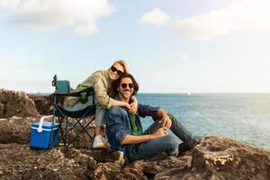 A couple relaxes on the rocky beach, with the woman playfully embracing the man. They enjoy their picnic, surrounded by water and a clear blue sky on a bright day. photo