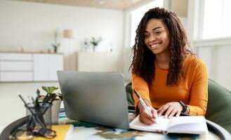 Smiling young African American woman sitting at desk working on laptop taking notes in notebook, happy millennial female studying online, watching webinar using computer and writing check list photo
