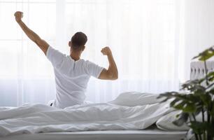 Rear View Of Young Man Stretching In Bed After Waking Up In The Morning, Unrecognizable Male Resting In Light Bedroom After Good Sleep, Looking At Window, Enjoying Start Of New Day, Copy Space photo