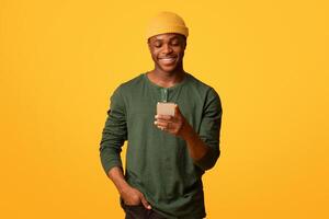 Cool App. Happy young black guy using smartphone, browsing new application or messaging with friends online on mobile phone, cheerful african man standing over yellow studio background, copy space photo