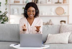 Online Tutoring Concept. Smiling black female tutor having call with student, giving language class by webcam, sitting on couch at home photo