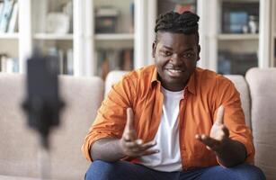Blogging concept. Young African American chuby guy blogger in casual clothes streaming online or recording tutorial for social media network, looking at the mobile phone on a tripod and smiling photo