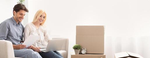 A young couple smiles while sitting on a couch, looking at documents and using a laptop. In the background, there are unpacked cardboard boxes and a small plant, indicating a recent move. photo