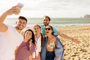 A group of five friends gathers on the sandy beach, smiling and taking selfies as the sun sets behind them, creating a joyful and lively atmosphere during their seaside outing. photo