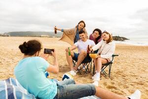 A group of friends sits on the sand, smiling and posing for a picture. They have snacks and drinks as they relax and enjoy their time together on a cloudy summer day. photo