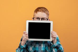 Ginger boy in eyeglasses hiding behind digital tablet with empty screen over yellow studio background. Cute redhead schoolboy showing pad, mockup, advertisement for cute application photo