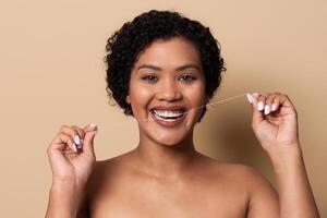 A woman with curly hair is smiling widely while holding dental floss. She demonstrates proper flossing technique in a relaxed, home environment with a neutral background. photo
