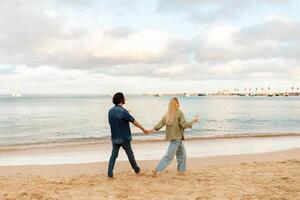 A couple strolls hand in hand along a sandy beach as the sun sets, with gentle ocean waves lapping at their feet. The sky features soft clouds and a warm glow of evening light. photo