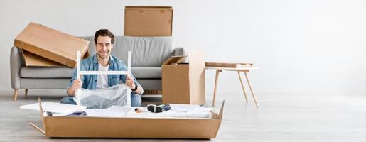 Positive emotions of person, moving to new house and assembling furniture with own hands. Young happy guy holding details on floor in living room interior with tools and cardboard boxes, empty space photo