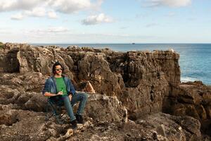 A man sits comfortably on a portable chair at a rocky coastal area, enjoying the ocean view. The clear sky and gentle waves create a peaceful atmosphere perfect for relaxation. photo