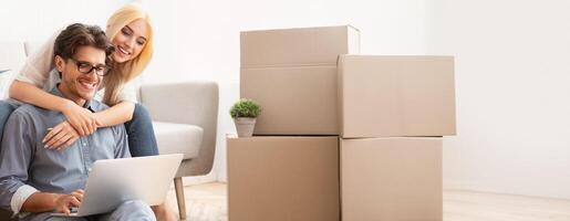 A young man types on a laptop while a woman embraces him from behind, surrounded by packed moving boxes in a bright living room. They appear happy and engaged, enjoying the moment, copy space photo