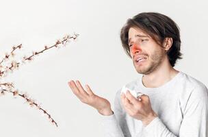 Seasonal allergies and health. Suffering man with red nose and napkin in hand looks at flowering plants, isolated on light background photo