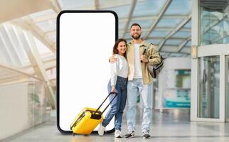 A young couple stands together in a bright airport terminal next to large phone with blank screen, one holding a yellow suitcase. photo