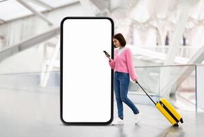 A young woman standing indoors at a modern location is using her phone while pulling a yellow suitcase. She is located next to a large blank digital advertising screen, suggesting a travel context. photo