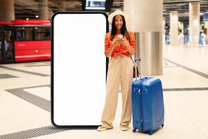 In a bustling airport, a young woman stands beside a large blank advertisement display while checking her phone. She holds a suitcase, appearing ready to embark on her journey. photo