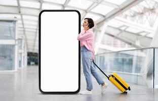 A traveler stands at the airport casually talking on a smartphone, pulling a yellow suitcase, passing by huge phone with blank screen photo